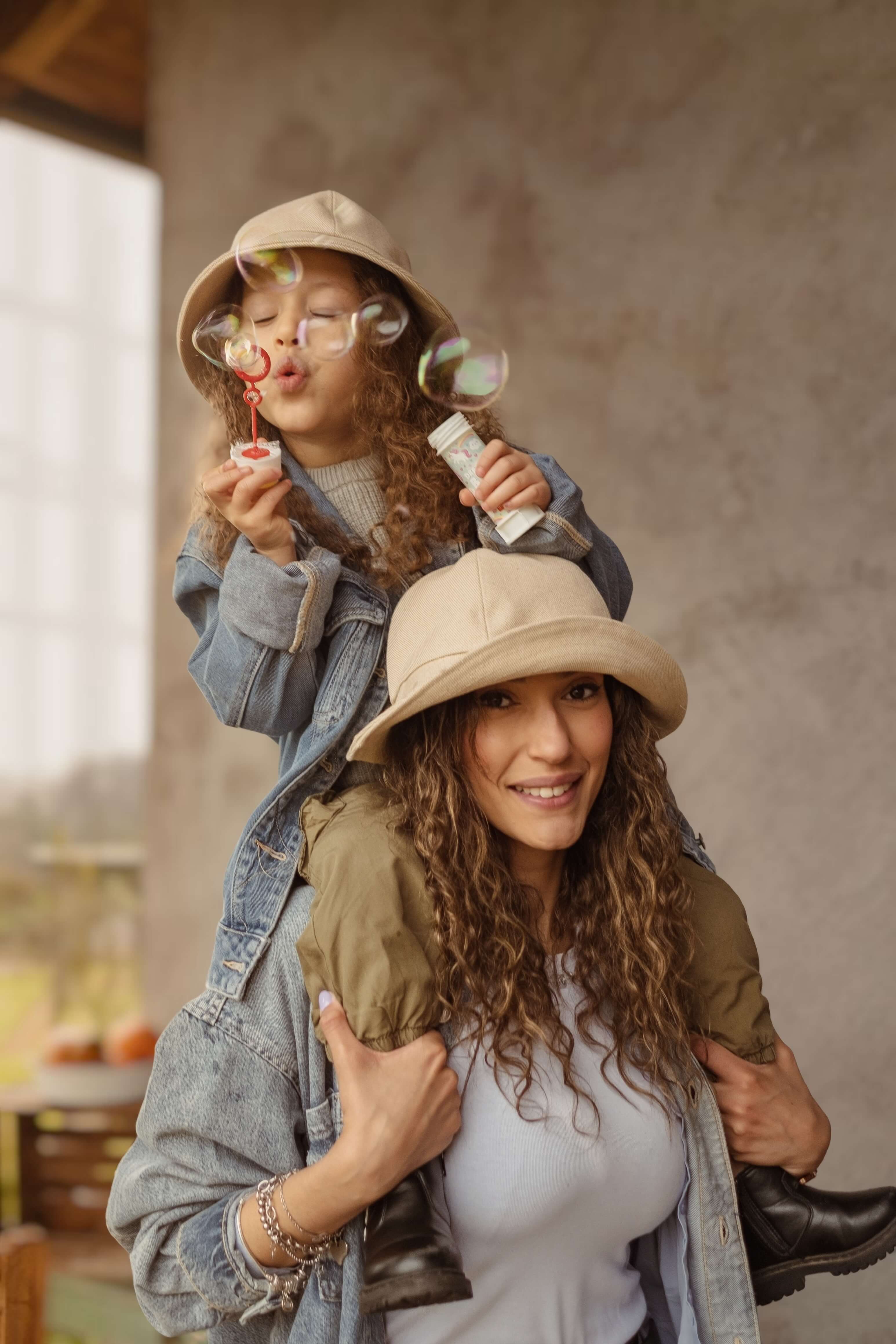 cappello bucket ies beige, coordinato mamma e figlia, immagine di una bimba in spalletta alla mamma mentre soffia le bolle di sapone